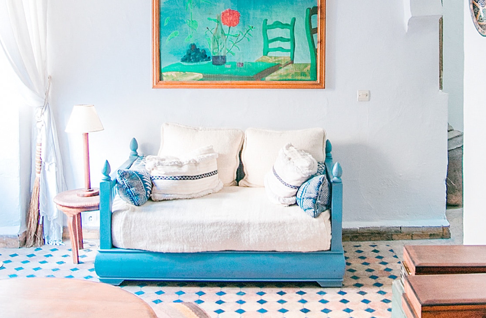 A Mediterranean-inspired living room with a cornflower blue daybed, a rose side table with light, and a still-life painting affixed on a white stucco wall.
