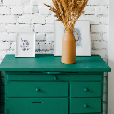 An emerald green chest of drawers topped with framed art and a vase of dried pampas grass.